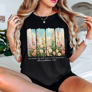 May include: Black t-shirt featuring a floral design with pink, yellow, and white flowers against a blue sky backdrop. The text "HIS MERCIES ARE NEW EVERY MORNING Lamentations 3:23" is printed below the floral design.