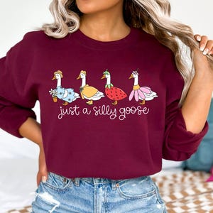 May include: Burgundy sweatshirt featuring the phrase "just a silly goose" in white script. The design includes four cartoon geese, each dressed in whimsical outfits, including a dress, cape, and strawberry costume. A fun and playful design.