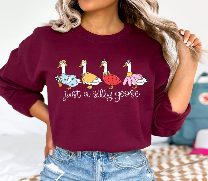 May include: Burgundy sweatshirt featuring the phrase "just a silly goose" in white script. The design includes four cartoon geese, each dressed in whimsical outfits, including a dress, cape, and strawberry costume. A fun and playful design.