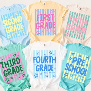 May include: Six short-sleeve t-shirts in light blue, yellow, cream, mint green, white, and teal. Each shirt features a vertical striped rectangular graphic with star accents and text reading First, Second, Third, Fourth, Fifth Grade, or Pre-School.