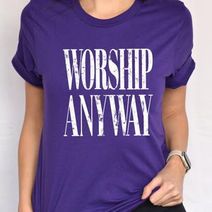 May include: A purple t-shirt with the words "WORSHIP ANYWAY" printed in large, distressed white letters. The shirt has short sleeves and a crew neck. The sleeves are rolled up.