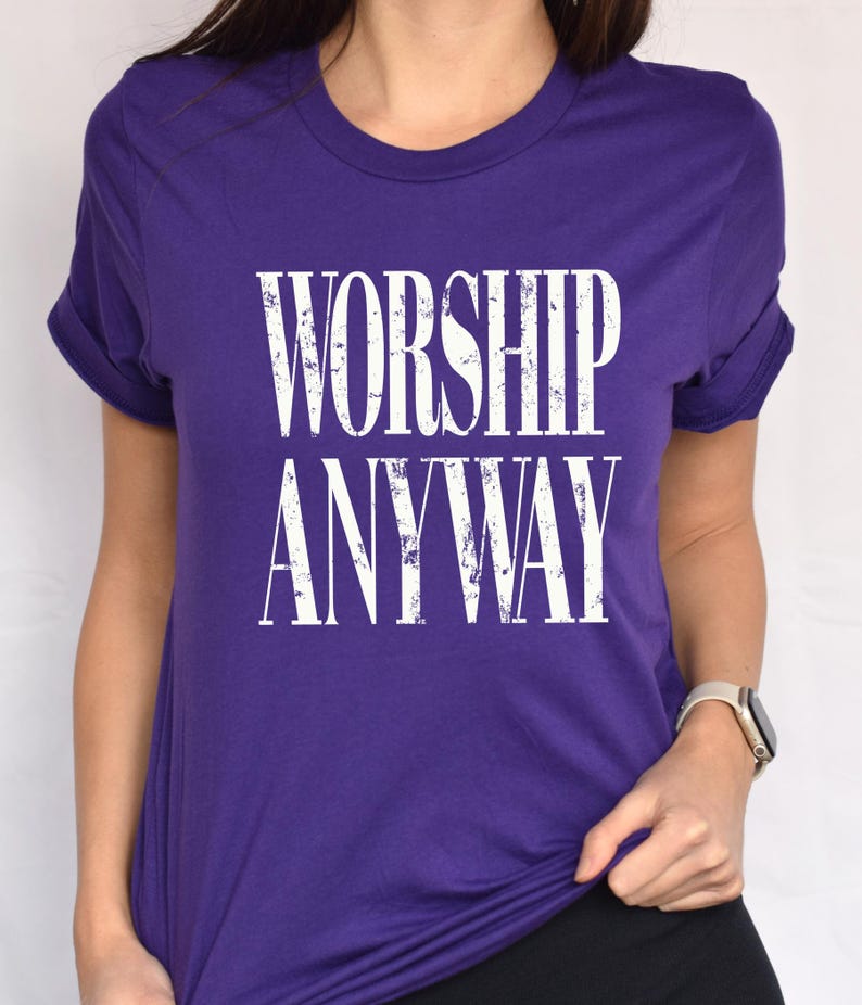 May include: A purple t-shirt with the words "WORSHIP ANYWAY" printed in large, distressed white letters. The shirt has short sleeves and a crew neck. The sleeves are rolled up.
