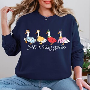 May include: Navy blue sweatshirt featuring a whimsical design of four geese, each adorned with unique accessories like a flower pot, cape, strawberry, and flower. The phrase "just a silly goose" is written below the geese.