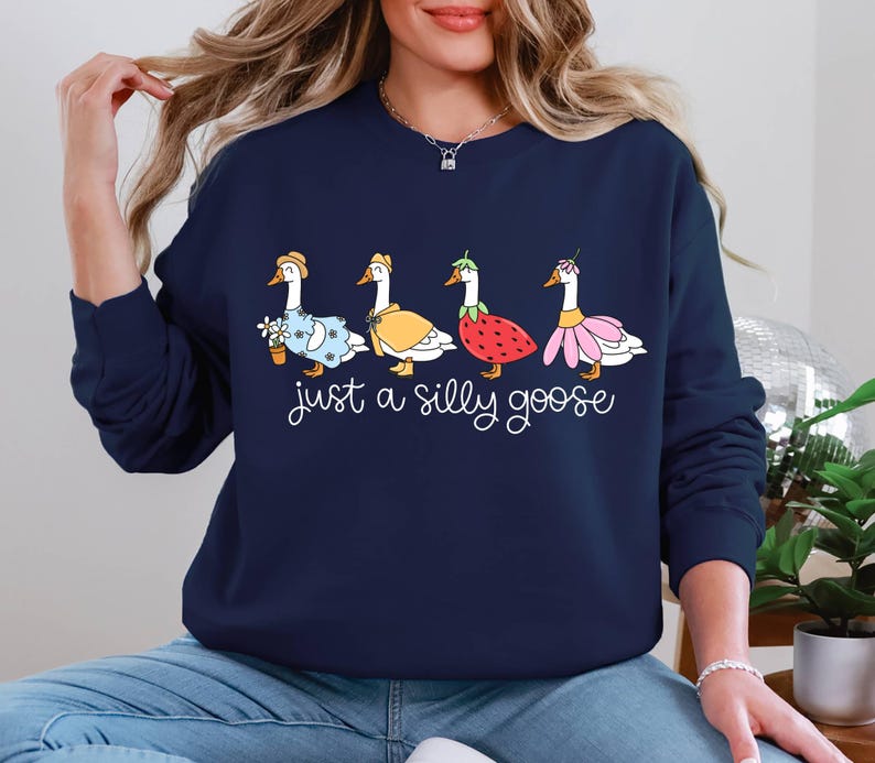 May include: Navy blue sweatshirt featuring a whimsical design of four geese, each adorned with unique accessories like a flower pot, cape, strawberry, and flower. The phrase "just a silly goose" is written below the geese.