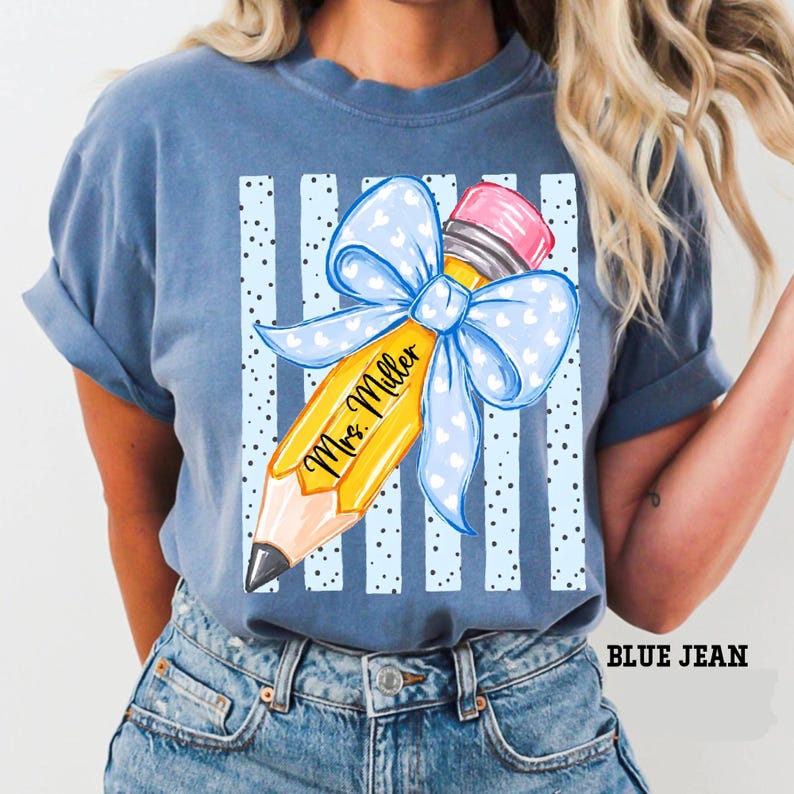 May include: A blue denim-colored t-shirt featuring a graphic of a yellow pencil with a pink eraser and a large light blue bow with white hearts. The pencil is printed with the text Mrs. Miller, set against a background of vertical stripes.