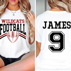 May include: White t-shirt with "WILDCATS FOOTBALL" in black and red lettering, and a football graphic. The back of the shirt displays "JAMES" above the number "9" in a black outlined box. A sporty apparel item.