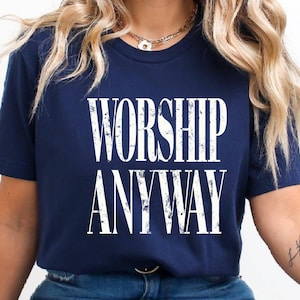 May include: Navy blue t-shirt with the words "WORSHIP ANYWAY" in distressed white lettering. The shirt has a crew neck and short sleeves. The text is centered on the front of the shirt.
