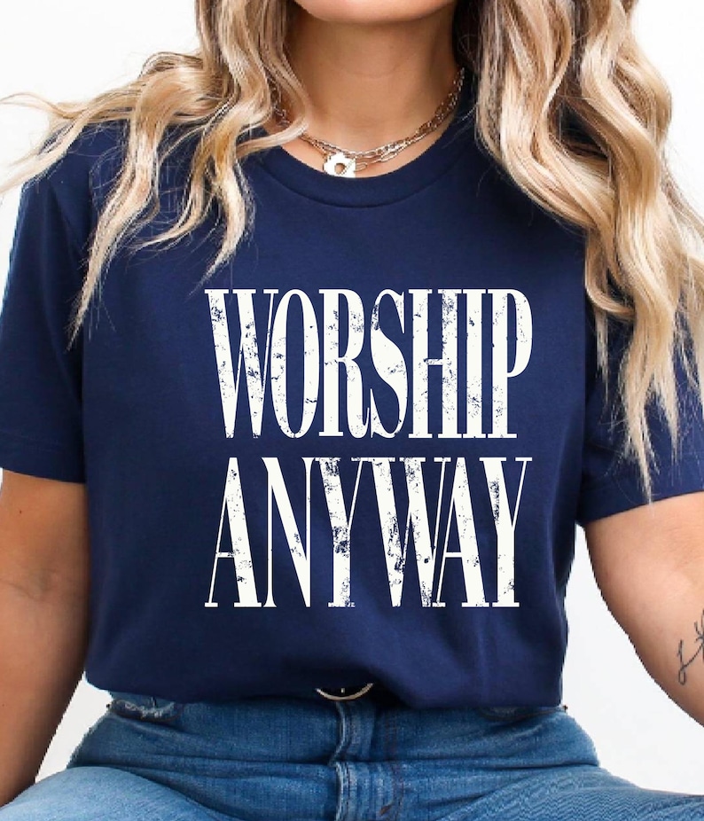 May include: Navy blue t-shirt with the words "WORSHIP ANYWAY" in distressed white lettering. The shirt has a crew neck and short sleeves. The text is centered on the front of the shirt.