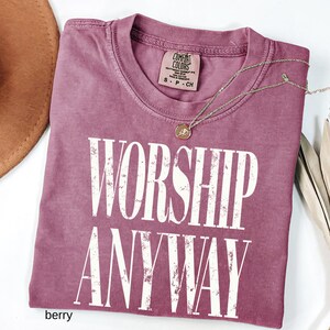 May include: Berry-colored t-shirt with the words "WORSHIP ANYWAY" in large white letters. The shirt has a crew neck and a small tag at the neckline. A gold-colored necklace with a pendant is draped across the shirt.