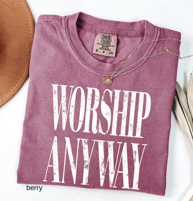 May include: Berry-colored t-shirt with the words "WORSHIP ANYWAY" in large white letters. The shirt has a crew neck and a small tag at the neckline. A gold-colored necklace with a pendant is draped across the shirt.