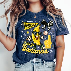 May include: Navy blue t-shirt with a cartoon banana wearing sunglasses and sneakers. The shirt features the text "BANANAS" on a banner and "Let's Go Bananas" in yellow, along with a baseball and a foam finger.