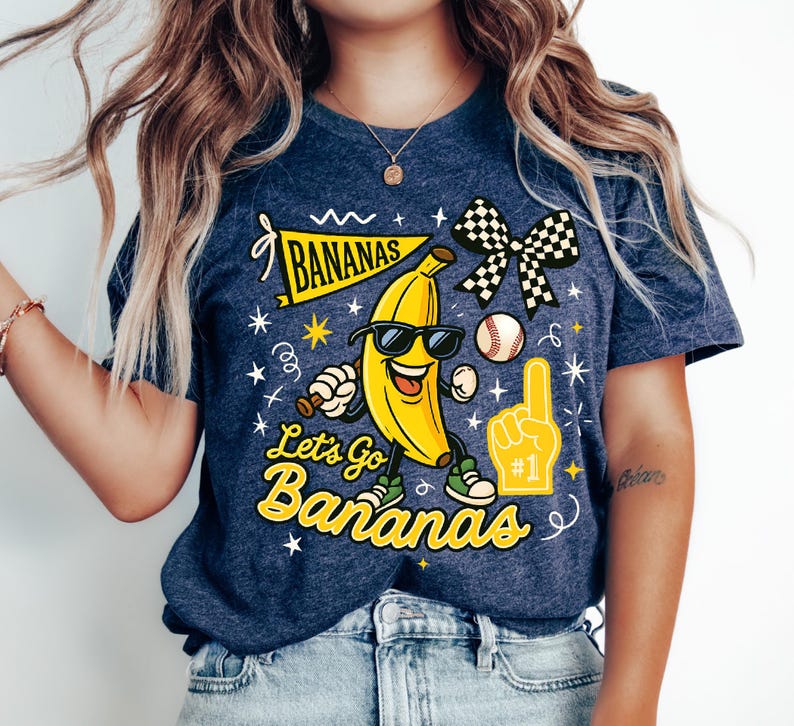 May include: Navy blue t-shirt with a cartoon banana wearing sunglasses and sneakers. The shirt features the text "BANANAS" on a banner and "Let's Go Bananas" in yellow, along with a baseball and a foam finger.