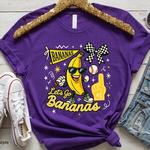 May include: A purple t-shirt featuring a cartoon banana wearing sunglasses, a baseball, and a checkered bow. The shirt has the text "BANANAS" and "Let's Go Bananas" in yellow. White sneakers are visible in the image.