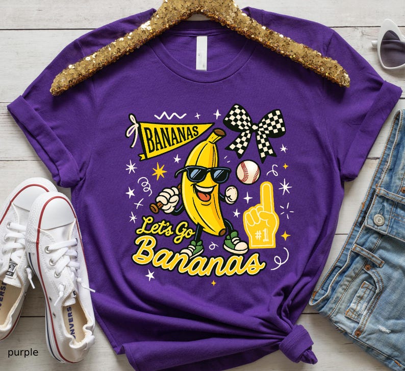 May include: A purple t-shirt featuring a cartoon banana wearing sunglasses, a baseball, and a checkered bow. The shirt has the text "BANANAS" and "Let's Go Bananas" in yellow. White sneakers are visible in the image.