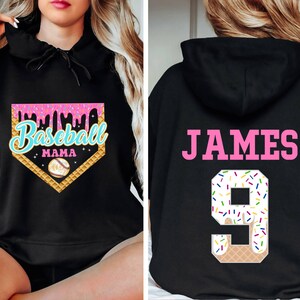 K&ouml;nnte beinhalten: Schwarzer Kapuzenpullover mit einem pinkfarbenen und blauen Baseball-Motiv auf der Vorderseite, auf dem "Baseball Mama" steht, und einer pinkfarbenen und wei&szlig;en Eiswaffel-Nummer 9 auf der R&uuml;ckseite mit dem Namen "JAMES" dar&uuml;ber.