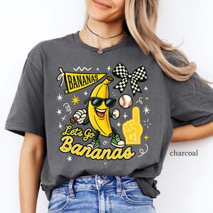 May include: Charcoal-colored t-shirt featuring a cartoon banana wearing sunglasses and sneakers, holding a baseball bat and a foam finger. The shirt has the text "Let's Go Bananas" and a banner that says "Bananas".