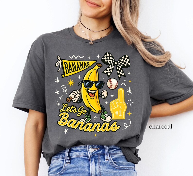 May include: Charcoal-colored t-shirt featuring a cartoon banana wearing sunglasses and sneakers, holding a baseball bat and a foam finger. The shirt has the text "Let's Go Bananas" and a banner that says "Bananas".