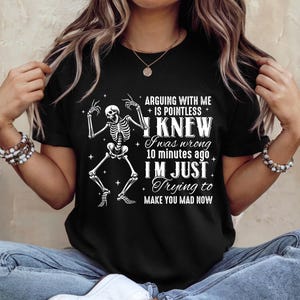 May include: Black t-shirt with a white skeleton graphic and the text "ARGUING WITH ME IS POINTLESS I KNEW I was wrong 10 minutes ago I'M JUST Trying to MAKE YOU MAD NOW". The shirt is a casual wear item.