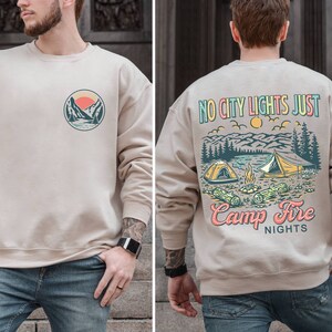 May include: A beige sweatshirt with a graphic of a mountain scene and the text "No City Lights Just Camp Fire Nights" on the back. The front of the sweatshirt has a smaller graphic of a mountain scene.