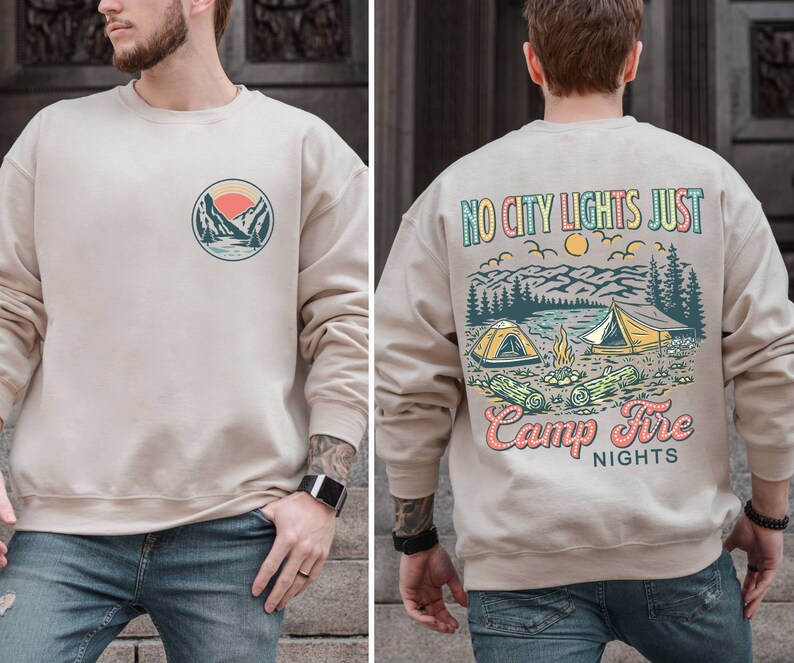 May include: A beige sweatshirt with a graphic of a mountain scene and the text "No City Lights Just Camp Fire Nights" on the back. The front of the sweatshirt has a smaller graphic of a mountain scene.