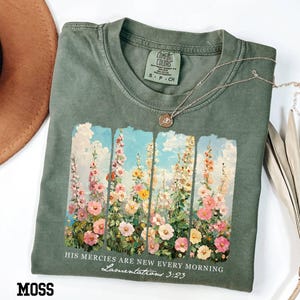 May include: Moss green t-shirt with a floral design featuring pink, yellow, and white flowers against a blue sky. The text "HIS MERCIES ARE NEW EVERY MORNING Lamentations 3:23" is printed below the floral image. A silver necklace with a pendant is on the shirt.