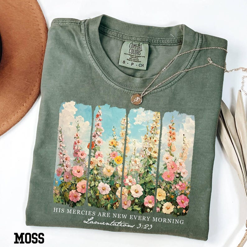 May include: Moss green t-shirt with a floral design featuring pink, yellow, and white flowers against a blue sky. The text "HIS MERCIES ARE NEW EVERY MORNING Lamentations 3:23" is printed below the floral image. A silver necklace with a pendant is on the shirt.