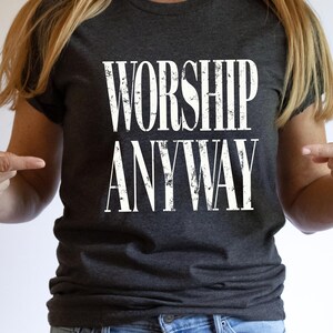 May include: Dark gray t-shirt with the words "WORSHIP ANYWAY" printed in distressed white lettering. The shirt is a crew neck style and is made of a soft material. The overall theme is religious and inspirational.
