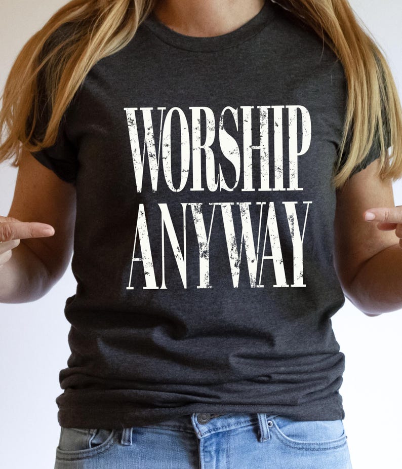 May include: Dark gray t-shirt with the words "WORSHIP ANYWAY" printed in distressed white lettering. The shirt is a crew neck style and is made of a soft material. The overall theme is religious and inspirational.