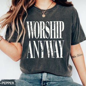 May include: Dark gray t-shirt with the words "WORSHIP ANYWAY" in distressed white lettering. The shirt is a crew neck style and is worn with blue jeans. A gold necklace is visible.