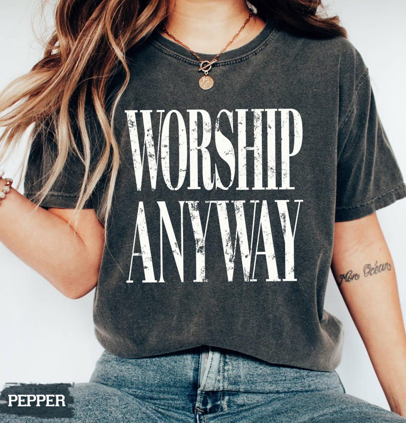 May include: Dark gray t-shirt with the words "WORSHIP ANYWAY" in distressed white lettering. The shirt is a crew neck style and is worn with blue jeans. A gold necklace is visible.