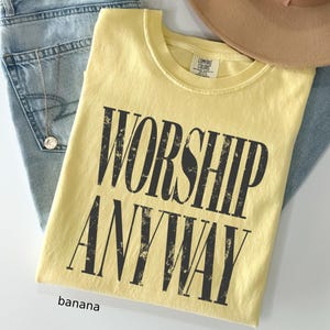 May include: A banana-colored t-shirt with the words "WORSHIP ANYWAY" printed in distressed black lettering. The shirt is folded and placed on top of blue jeans and a tan hat.