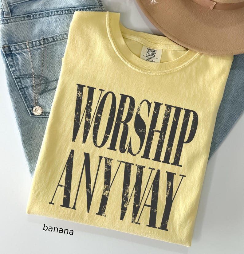 May include: A banana-colored t-shirt with the words "WORSHIP ANYWAY" printed in distressed black lettering. The shirt is folded and placed on top of blue jeans and a tan hat.