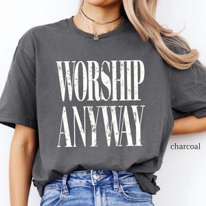 May include: Charcoal-colored t-shirt with the words "WORSHIP ANYWAY" in a distressed white font. The shirt is a relaxed fit and is paired with blue jeans. The word "charcoal" is printed in the lower right corner.