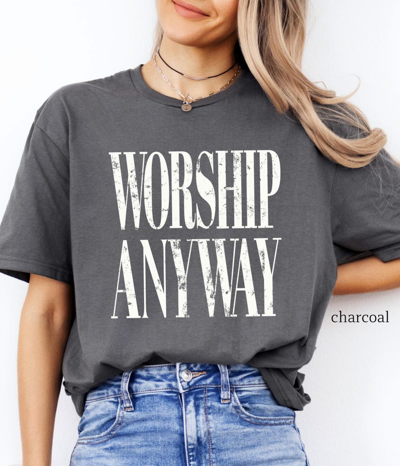 May include: Charcoal-colored t-shirt with the words "WORSHIP ANYWAY" in a distressed white font. The shirt is a relaxed fit and is paired with blue jeans. The word "charcoal" is printed in the lower right corner.