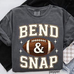 May include: A dark gray sweatshirt with the words "BEND & SNAP" in white and tan block letters. A brown football graphic is centered below the text, with white star accents on either side. The word "pepper" is at the bottom.