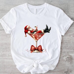 May include: White t-shirt with a graphic of Santa Claus relaxing in a martini glass with a red bow on the stem.