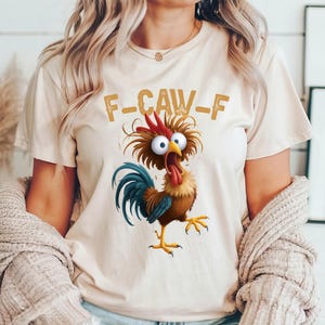 May include: Beige t-shirt featuring a cartoon rooster with a red comb, blue tail feathers, and yellow feet. The rooster has wide eyes and a long red tongue. The text "F-CAW-F" is printed above the rooster in a brown, distressed font.