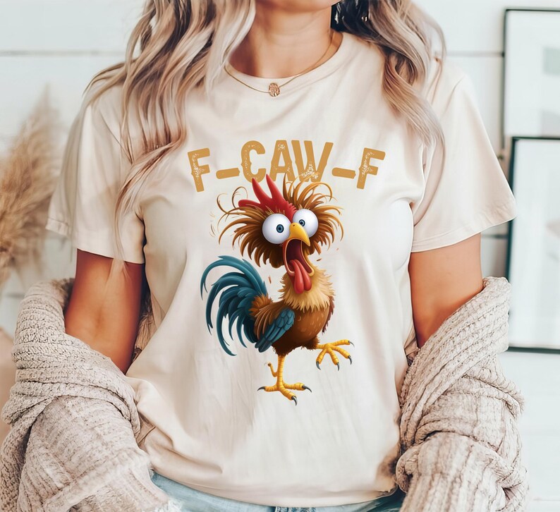 May include: Beige t-shirt featuring a cartoon rooster with a red comb, blue tail feathers, and yellow feet. The rooster has wide eyes and a long red tongue. The text "F-CAW-F" is printed above the rooster in a brown, distressed font.