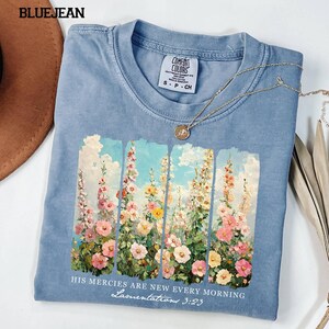 May include: A light blue t-shirt with a floral design featuring pink, yellow, and white flowers against a blue sky. The text "HIS MERCIES ARE NEW EVERY MORNING Lamentations 3:23" is printed below the floral design. A silver necklace is draped across the shirt.