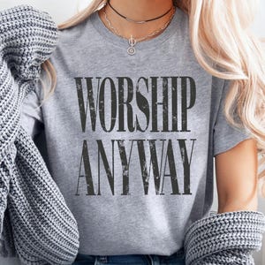 May include: A gray t-shirt with the words "WORSHIP ANYWAY" printed in a distressed black font. The shirt is short-sleeved and has a crew neck. The image shows the shirt being worn with a necklace and a gray sweater draped over the shoulders.