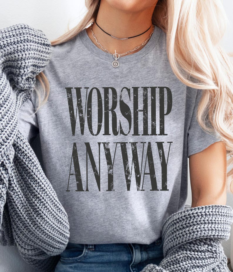 May include: A gray t-shirt with the words "WORSHIP ANYWAY" printed in a distressed black font. The shirt is short-sleeved and has a crew neck. The image shows the shirt being worn with a necklace and a gray sweater draped over the shoulders.