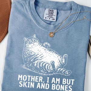 以下が含まれることがあります： 白い寝そべり猫のグラフィックと「MOTHER, I AM BUT SKIN AND BONES」の文字が入った、ブルーデニムカラーのTシャツ。小さなペンダントが付いたゴールドカラーのネックレスがシャツの上にあります。シャツにはComfort Colorsのタグが付いています。