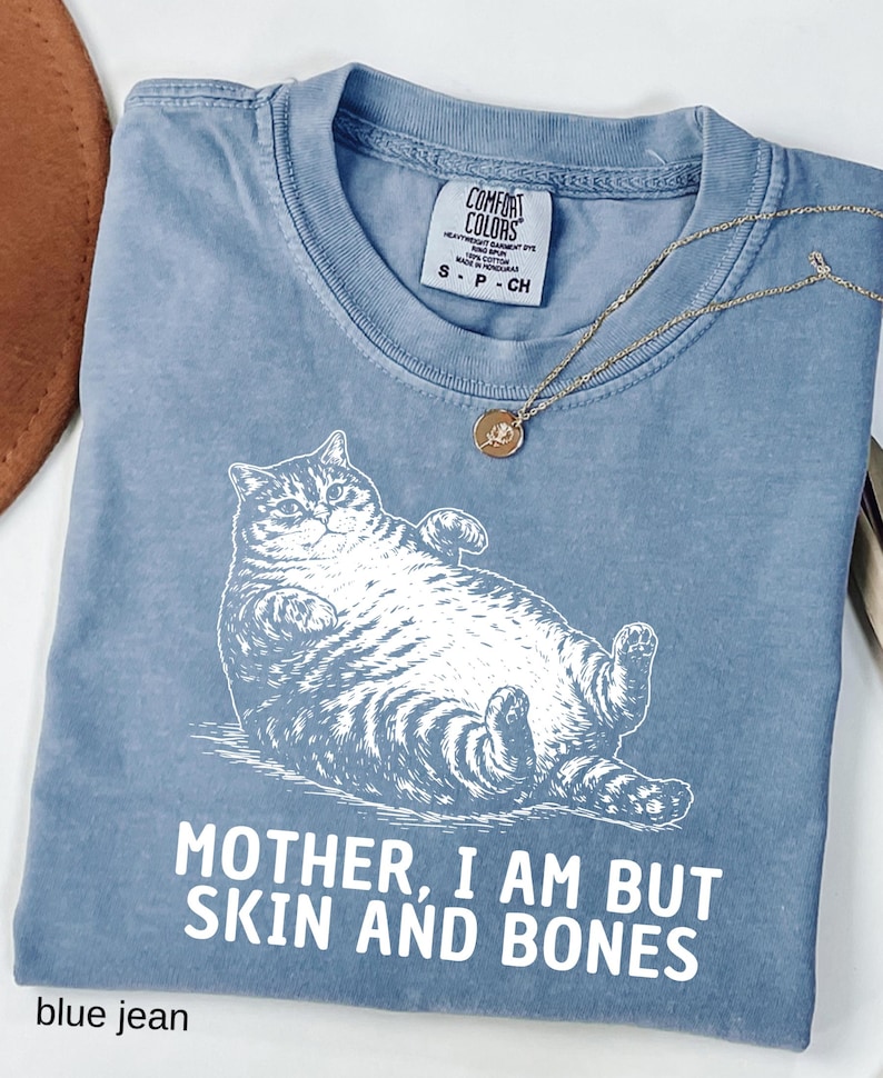 以下が含まれることがあります： 白い寝そべり猫のグラフィックと「MOTHER, I AM BUT SKIN AND BONES」の文字が入った、ブルーデニムカラーのTシャツ。小さなペンダントが付いたゴールドカラーのネックレスがシャツの上にあります。シャツにはComfort Colorsのタグが付いています。