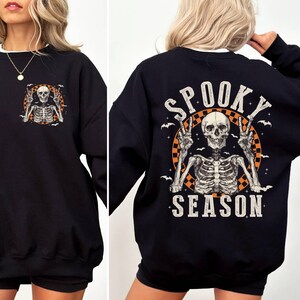 Puede incluir: Sudadera negra con un gr&aacute;fico de esqueleto y el texto "Spooky Season" impreso en la espalda. El esqueleto est&aacute; levantando dos signos de paz y est&aacute; rodeado de murci&eacute;lagos.