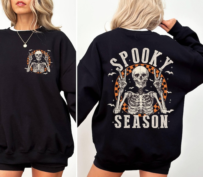 Puede incluir: Sudadera negra con un gr&aacute;fico de esqueleto y el texto "Spooky Season" impreso en la espalda. El esqueleto est&aacute; levantando dos signos de paz y est&aacute; rodeado de murci&eacute;lagos.
