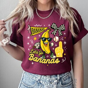 May include: Maroon t-shirt with a graphic of a smiling banana wearing sunglasses, sneakers, and holding a baseball bat. The text "BANANAS" and "Let's Go Bananas" are also on the shirt, along with a baseball, foam finger, and checkered bow.