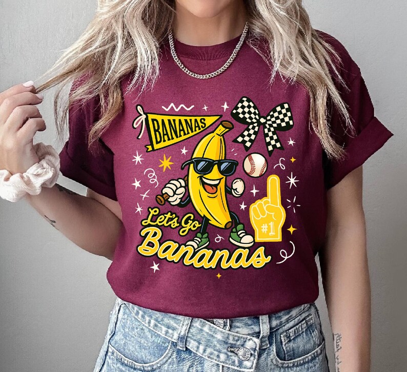 May include: Maroon t-shirt with a graphic of a smiling banana wearing sunglasses, sneakers, and holding a baseball bat. The text "BANANAS" and "Let's Go Bananas" are also on the shirt, along with a baseball, foam finger, and checkered bow.