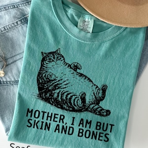 以下が含まれることがあります： 猫の白黒イラストがプリントされた、シーフォームグリーンのTシャツ。「MOTHER, I AM BUT SKIN AND BONES」の文字が猫の下にプリントされています。柔らかい素材で作られたシャツです。