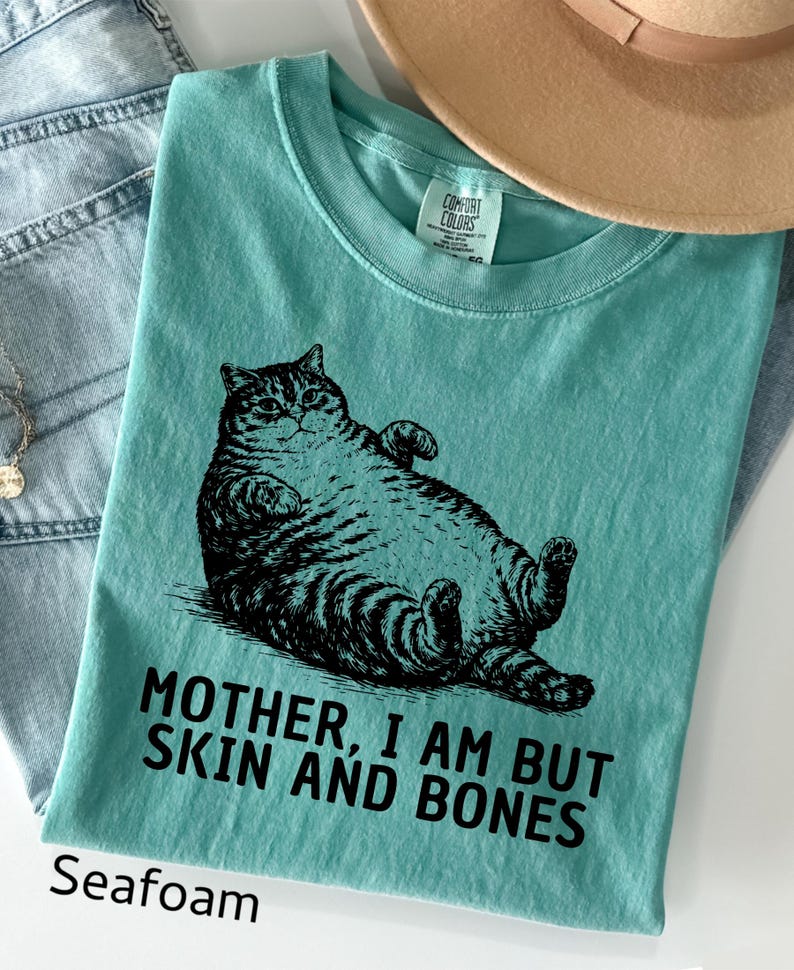 以下が含まれることがあります： 猫の白黒イラストがプリントされた、シーフォームグリーンのTシャツ。「MOTHER, I AM BUT SKIN AND BONES」の文字が猫の下にプリントされています。柔らかい素材で作られたシャツです。