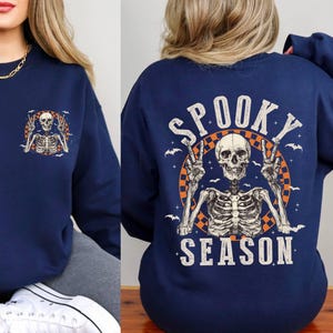 Puede incluir: Sudadera azul marino con un gr&aacute;fico de esqueleto y el texto "Spooky Season" en blanco y naranja. El esqueleto lleva un collar con un signo de paz y tiene los brazos levantados en un gesto de paz.